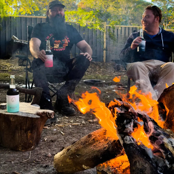 Two men sitting by a campfire outdoors, drinking from Solkatt Designs insulated stubby coolers — perfect for keeping drinks cold on chilly nights.